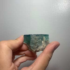 May include: A green fluorite crystal with white inclusions. The crystal is rough and has a natural, unpolished look.