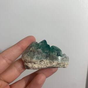 May include: A green fluorite crystal cluster with a rough, natural surface. The crystal is a light green colour with some white inclusions.