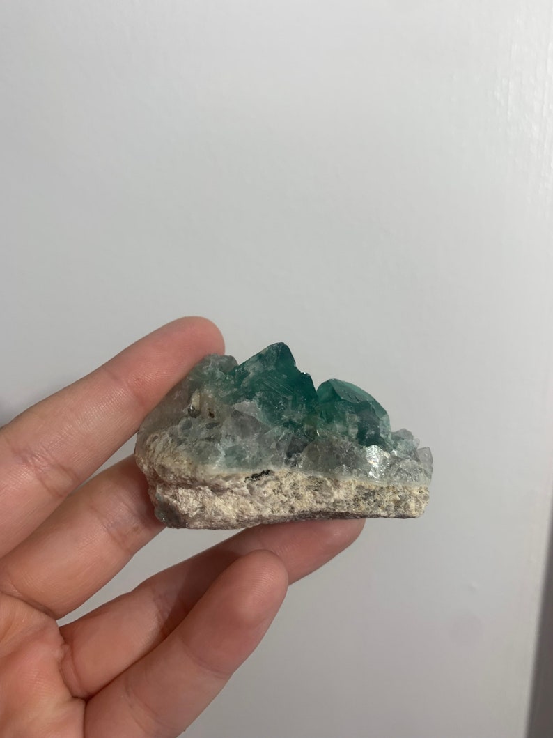May include: A green fluorite crystal cluster with a rough, natural surface. The crystal is a light green colour with some white inclusions.