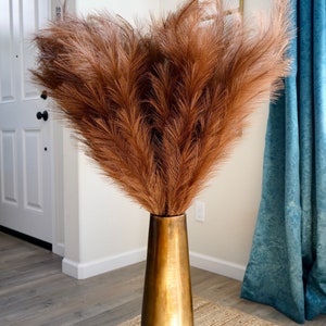 May include: A bouquet of dried pampas grass in a tall, gold vase. The vase is on a woven rug in front of a white door and a blue curtain.