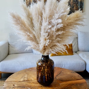 May include: A vase with a brown and black leopard print design filled with dried pampas grass sits on a wooden coffee table with a unique, irregular shape.