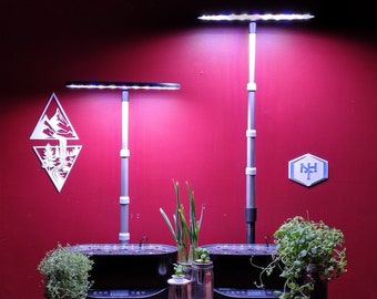 May include: Two black hydroponic gardening systems with white LED grow lights on adjustable poles. The lights are on and illuminating the plants. The systems are on a wooden table with a red wall in the background.