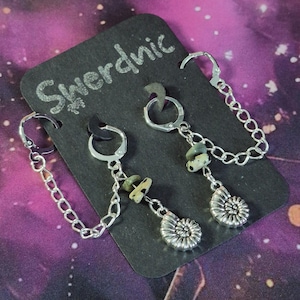May include: Silver earrings featuring a chain, small green stones, and a spiral seashell charm. The earrings are presented on a black card with the word "Swerdnic" in silver. The background is a purple and black galaxy design.