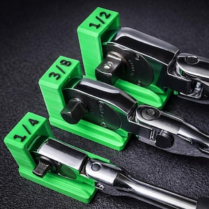 May include: Three chrome ratchet wrenches with green holders labeled 1/4, 3/8, and 1/2. The wrenches are arranged on a dark surface, showcasing their shiny metal finish and the bright green of the holders. The tools are for mechanical work.