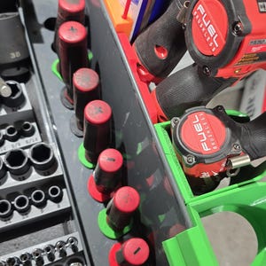 May include: A gray tool organizer tray containing a row of black screwdrivers with red caps marked with plus and minus symbols, a set of black metal socket bits, and two red Milwaukee Fuel Brushless power tools stored in green plastic holders.