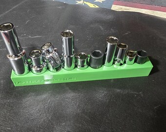 Puede incluir: Un juego de llaves de vaso cromadas de varios tamaños, dispuestas sobre un soporte verde brillante. Las llaves de vaso son cilíndricas con aberturas hexagonales. El soporte tiene el texto "POWERED BY DESIGNS" impreso.