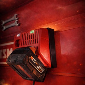 May include: A red and black battery charger mounted on a red metal surface, featuring a black M18 Redlithium XC5.0 battery inserted into the charging port. The charger has vertical cooling vents and a black power cord extending downward.