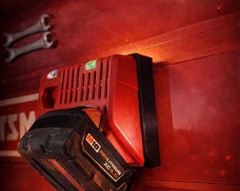 Puede incluir: Una batería Milwaukee M18 Redlithium XC 5.0 roja y negra montada en una estación de carga roja. La batería tiene una carcasa negra con detalles rojos y texto blanco. El cargador está fijado a una superficie metálica roja con dos llaves.