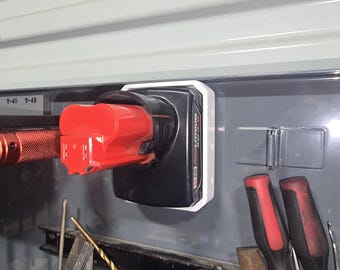 Puede incluir: Una batería roja y negra para herramientas eléctricas está enchufada a un cargador blanco y negro montado en una pared gris. Se ven otras herramientas, como una llave inglesa y destornilladores.