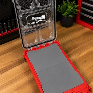 Könnte beinhalten: Ein roter und durchsichtiger Kunststoff-Aufbewahrungskoffer mit Klappdeckel. Der Deckel verfügt über ein Sichtfenster mit Wabenmuster und das Milwaukee Packout-Logo. Die Basis enthält zwei flache, graue Schaumstoffeinsätze zur Werkzeugaufbewahrung.