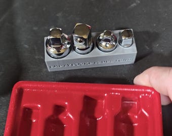 Puede incluir: Un juego de llaves de vaso cromadas en un soporte gris, con el texto "EMPTY POCKETS DESIGNS". Una bandeja de plástico roja contiene las llaves de vaso. La imagen está tomada desde un ángulo ligeramente elevado.