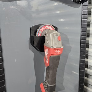 May include: A black plastic wall-mounted holder secures a red and gray Milwaukee angle grinder with a Bauer flap disc attached. The tool is positioned vertically against a gray metal surface, featuring a black handle and a red battery base.