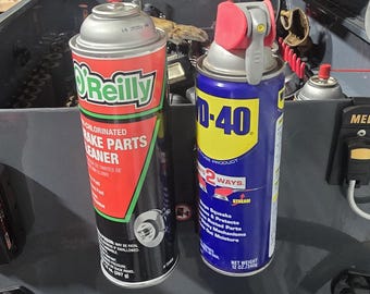 Puede incluir: Dos botes de aerosol, uno con la etiqueta "O'Reilly Chlorinated Brake Parts Cleaner" y el otro "WD-40". El bote de limpiador de frenos es rojo, negro y verde. El bote de WD-40 es azul y amarillo. Ambos botes están sobre una superficie gris oscura.