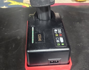 Puede incluir: Cargador de batería negro y rojo con una batería negra adjunta. El cargador tiene un puerto de alimentación e indicadores luminosos. El cargador está sobre una superficie negra. El cargador tiene el número 1065946 impreso.