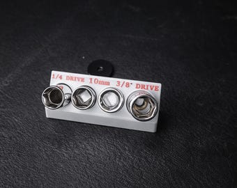 Puede incluir: Un juego de cuatro llaves de vaso cromadas en un soporte blanco, con el texto "1/4 DRIVE 10mm 3/8" DRIVE" en rojo. Una arandela negra está encima del juego de llaves de vaso. Las llaves de vaso son para trabajos mecánicos.
