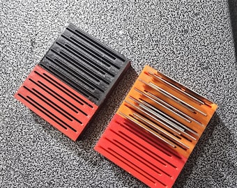 Puede incluir: Dos organizadores rectangulares con ranuras paralelas. El organizador izquierdo es negro y rojo con ranuras vacías. El organizador derecho es naranja y rojo, y contiene varias agujas o alfileres metálicos, finos y cilíndricos.