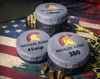 Puede incluir: Contenedores de almacenamiento de munición apilables con tapas de rosca. Los contenedores grises tienen el texto "MOLON LABE" y las marcas de calibre: 10mm, 45acp y 380. La imagen incluye balas y el texto "APILABLE", "COMPACTO Y PORTÁTIL" y "VETERAN-OWNED".