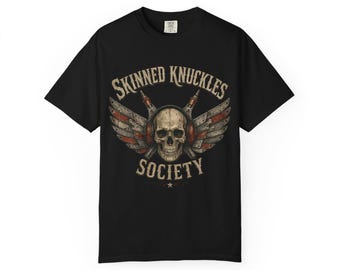 Puede incluir: Camiseta negra con un diseño gráfico vintage. El diseño presenta una calavera con auriculares, alas y el texto "SKINNED KNUCKLES SOCIETY". El gráfico utiliza tonos rojos, blancos y marrones.