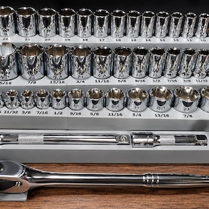 May include: A comprehensive chrome socket set organized on gray tiered trays. The collection includes various sizes labeled with imperial measurements, a ratchet handle marked 3/8, extension bars, and a universal joint, all featuring a polished finish.