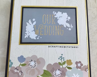 May include: A white and gold scrapbook with a woodgrain background. The front cover features a gray rectangle with the text "OUR WEDDING" in gold lettering and white floral accents. The bottom of the cover has a floral design with pink, blue, and green flowers.