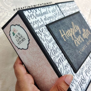 May include: A black and gray scrapbook with a pink and white patterned cover. The cover has the text "Our Love Story" on it. The scrapbook is open to reveal a page with the text "Happily Ever After Starts Here".