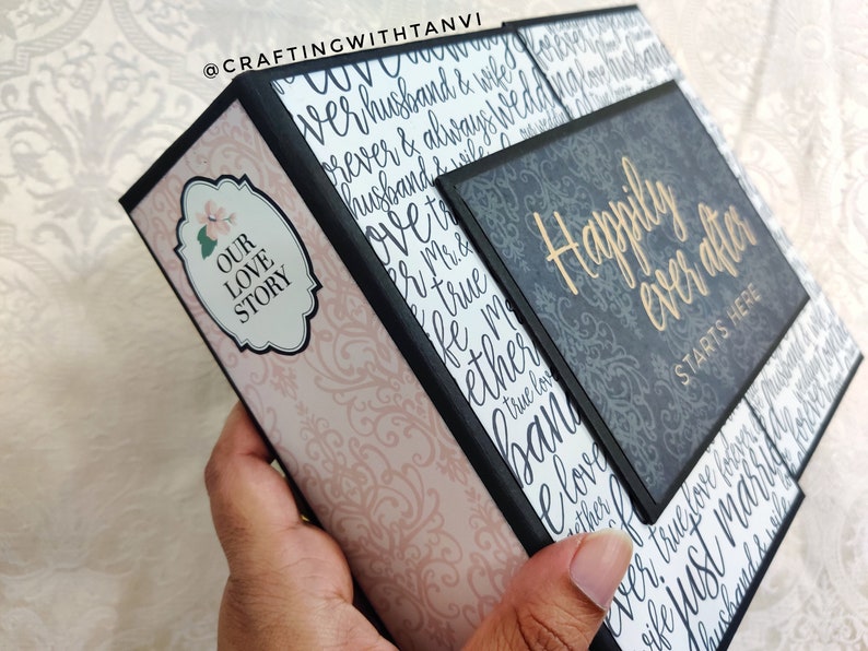 May include: A black and gray scrapbook with a pink and white patterned cover. The cover has the text "Our Love Story" on it. The scrapbook is open to reveal a page with the text "Happily Ever After Starts Here".