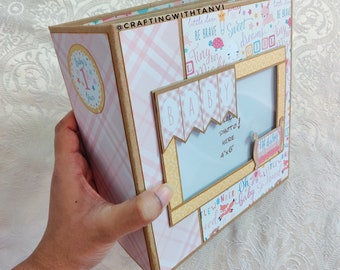 May include: A pink and brown scrapbook with a photo frame and a baby theme. The scrapbook has a pink and white checkered pattern and features the words "Baby", "Be Brave", "Sweet Dreams", "Tiny", "Little Deer", and "Oh So Loved".