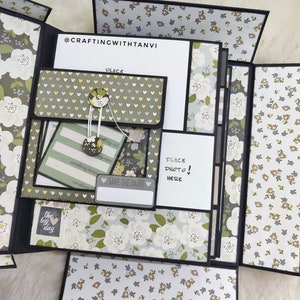 May include: A multi-page scrapbook with a floral pattern and a white background. The scrapbook has a pocket on the front with a button closure. The text "SAVE THE DATE!" is written on a white tag. The text "PLACE PHOTO HERE!" is written on a white card.