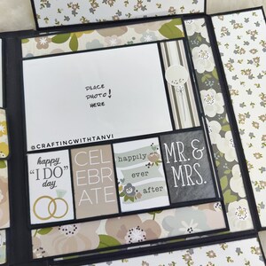 May include: A black scrapbook with a floral pattern on the cover. The scrapbook is open to reveal a white card with the words "Place Photo Here!" written on it. The scrapbook also has several other cards with text such as "Happy "I Do" Day", "Celebrate", "Happily Ever After", and "Mr. & Mrs.".