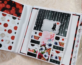 May include: A white scrapbook with red and black patterned pages. The scrapbook is open to reveal a page with a black and white silhouette of a couple dancing with a red heart above them. The page also features a floral design with pink, red, and white flowers.