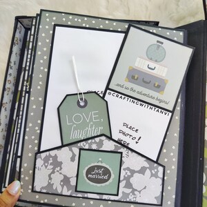 May include: A grey scrapbook with a floral pattern and black accents. The scrapbook has a white card with the text "Place Photo Here!" and a green tag with the text "Just Married". The scrapbook also has a green tag with the text "Love, Laughter" and a white card with the text "and so the adventure begins! @CRAFTINGWITHTANVI".