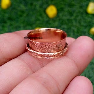 May include: A copper-coloured spinner ring featuring an embossed floral pattern on the wide central band and a textured geometric design on the thinner outer band. The text shinegemsjewel925 is visible on the inner surface of the ring.