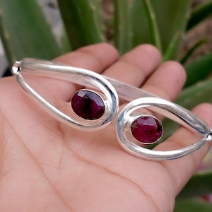 May include: A silver-toned cuff bracelet featuring an open, looped design on each end. Each loop holds a single, polished, oval-shaped deep red gemstone. The metal has a smooth, reflective finish, and the bracelet is constructed with a hinged mechanism.