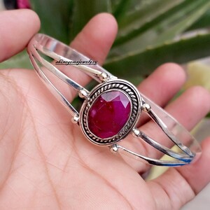 May include: A silver cuff bracelet featuring a central oval-shaped faceted deep pink gemstone set in a twisted metal border. The band consists of three parallel silver wires that converge at the setting. The text shinegemsjewel925 is visible.