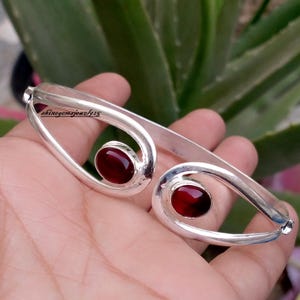 May include: A silver cuff bracelet featuring two open, teardrop-shaped ends, each set with a smooth, oval-shaped deep red gemstone. The metal has a polished finish, and the text shinegemsjewel925 is visible on the upper left side of the frame.