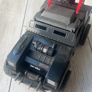 Pode incluir: Caminhão de brinquedo preto com uma pega vermelha no topo. O caminhão tem uma grelha preta e um motor preto com um tubo de escape prateado. O caminhão tem um pára-choques preto com um gancho prateado. O caminhão tem uma placa de matrícula preta que diz "BW-XG-4".