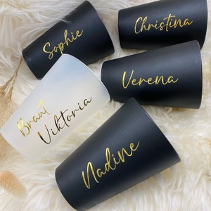 Könnte beinhalten: Fünf personalisierte schwarze und mattierte Becher mit goldfarbenen Buchstaben. Die Namen Sophie, Christina, Verena, Nadine und Brant Viktoria sind auf den Bechern abgebildet. Die Becher sind auf einer weißen, flauschigen Oberfläche angeordnet.