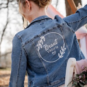 Könnte beinhalten: Eine blaue Jeansjacke mit einem weißen Kreisdesign auf der Rückseite. Das Design enthält das Wort "Braut" und den Namen "Viktoria" mit einem Herzsymbol.