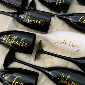 Könnte beinhalten: Schwarze und weiße Champagnerflöten mit goldener Beschriftung. Die Flöten sind mit Namen verziert, darunter "Anna", "Steffi", "Vivien", "Nathalie", "Bride Vany", "Isa" und "Johanna".