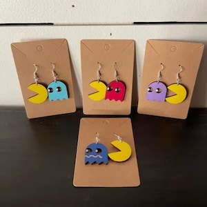 May include: Four pairs of Pac-Man and ghost earrings. The earrings are made of wood and painted in different colors. The Pac-Man earrings are yellow and the ghost earrings are blue, red, purple, and blue.