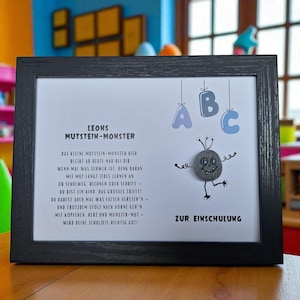 Könnte beinhalten: Ein gerahmter Druck mit einem schwarzen Holzrahmen. Der Druck zeigt Text und ein Cartoon-Steinmonster mit dem Text "LEONS MUTSTEIN-MONSTER" und "ZUR EINSCHULUNG". Der Hintergrund ist weiß mit blauen und grauen Akzenten.