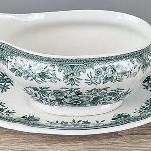 Könnte beinhalten: Eine weiße Keramik-Sauciere mit passender ovaler Untertasse, verziert mit einem dunkelgrünen Blumenmuster. Die Sauciere hat einen geschwungenen Ausguss und einen seitlichen Griff, während die Untertasse einen gewellten Rand mit botanischen Motiven aufweist.