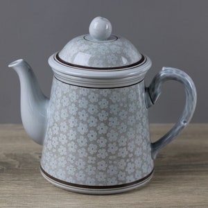 Könnte beinhalten: Eine graue Teekanne aus Keramik mit einem weißen Blumenmuster und einem braunen Rand. Die Teekanne hat einen Deckel und einen Griff.