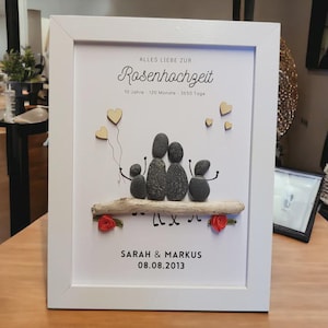 Könnte beinhalten: Weiß gerahmtes Kunstwerk mit einer Familie aus fünf Personen, dargestellt mit glatten, dunkelgrauen Steinen. Das Kunstwerk enthält Holzherzen, rote Rosen und die Namen "Sarah & Markus" mit einem Datum.