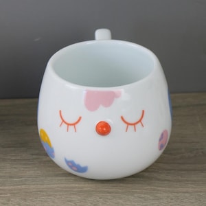 Könnte beinhalten: Weißer Keramikbecher mit einem skurrilen Design. Der Becher hat geschlossene orangefarbene Wimpern, eine kleine orangefarbene Nase und farbenfrohe Akzente in Rosa, Blau und Gelb. Der Becher hat einen kleinen Griff und eine abgerundete Form.