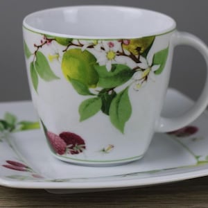 Könnte beinhalten: Eine weiße Kaffeetasse aus Keramik mit passender quadratischer Untertasse, verziert mit einem botanischen Druck aus grünen Limetten, kleinen roten Beeren und weißen Blüten mit grünen Blättern. Die Tasse hat einen runden Henkel und eine glatte Oberfläche.