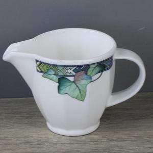 Peut inclure: Un pot à lait en céramique blanche doté d'un bec verseur et d'une anse latérale. L'extérieur est orné d'une bande horizontale aux motifs géométriques bleus, verts et blancs, agrémentée d'un groupe de feuilles de lierre vertes et violettes.
