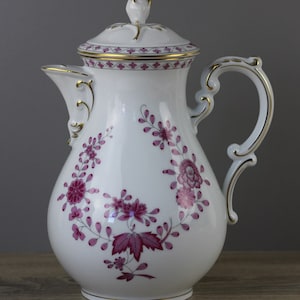 Peut inclure: Cafetière en porcelaine blanche avec un motif floral rose et des accents dorés. La cafetière a un couvercle et une poignée incurvée.
