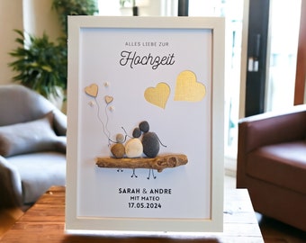 May include: A white framed picture with a white background. The picture features a family of three made of stones sitting on a branch. The family is holding hands and there are three hearts floating above them. The text "Alles Liebe zur Hochzeit" is at the top of the picture. Below the family is the text "Sarah & Andre mit Mateo 17.05.2024".