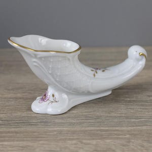 May include: A white porcelain cornucopia-shaped vase with gold trim and delicate floral accents. The vase has a curved, horn-like shape with a detailed textured surface and a small base. The design includes purple flowers and gold detailing.